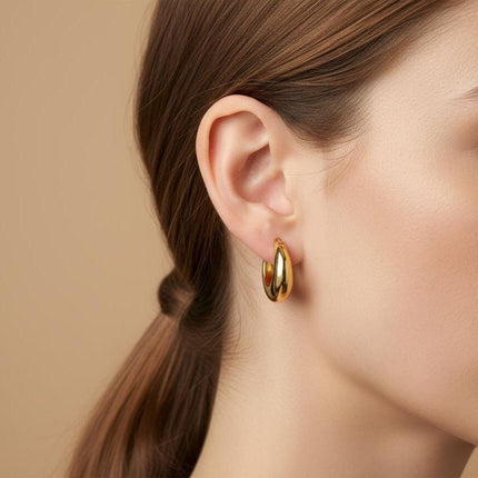 May include: A close-up of a woman's ear with a gold hoop earring. The earring is a thick, rounded design with a polished finish. The woman has brown hair pulled back in a ponytail, and her skin is fair with a neutral background.
