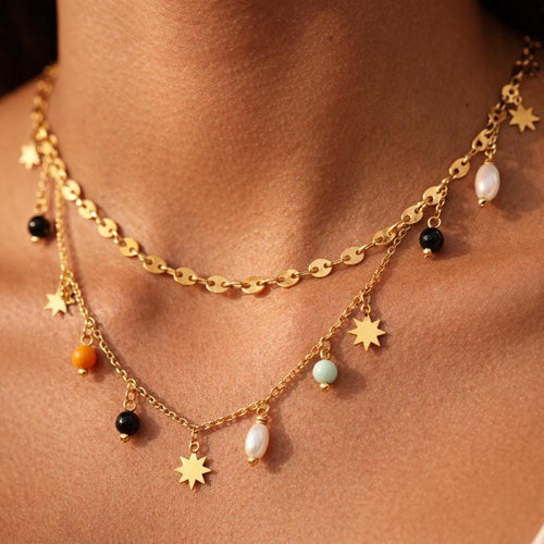 Close-up of a tanned neck wearing a layered gold necklace—one disc chain and one charm chain with tiny sun/star charms and small bead drops in black, orange, mint, and pearl-like white, in warm natural lighting.