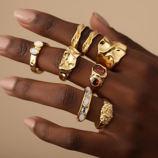 8 piece gold ring set featuring chunky molten textured bands, an amber gemstone statement ring, pearl and opal inlay rings, a wide sculptural gold band, and a twisted braided ring — arranged flat lay on warm beige surface
