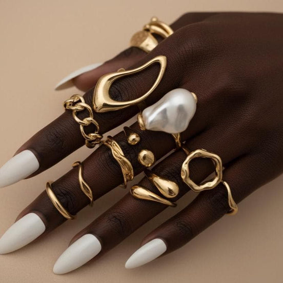12 piece gold ring set featuring a baroque pearl statement ring, hollow sculptural oval ring, gold chain link band, molten wavy stacking rings, teardrop ring, ball-end cuff, and twisted open bands — all worn stacked across a tanned woman's hand with long white almond nails