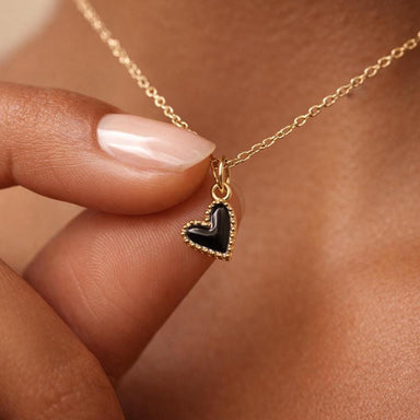 Tanned woman's hand pinching a small black enamel heart pendant necklace with gold beaded frame border on a delicate gold chain, dainty black heart charm necklace for women, minimalist gold jewelry close-up