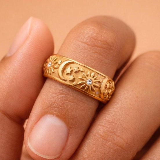 Close-up of a tanned hand wearing a gold celestial band ring with sunburst details, a crescent moon and stars, and small clear stones, photographed on a soft warm peach/beige background.