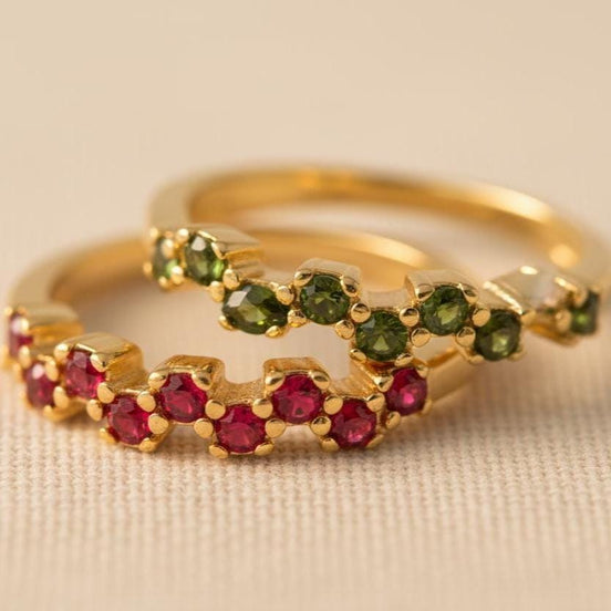 Two dainty gold open band rings with scattered round CZ gemstones in blue, teal, green, and red arranged in a diagonal display on a soft beige ceramic tray, delicate birthstone stacking rings for women, minimalist gold jewelry